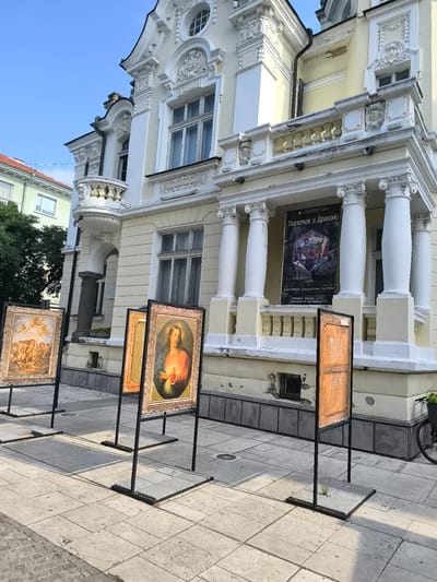 This image depicts an outdoor art exhibition in front of an ornate, light-yellow historical building, likely Uzunova House at Tsar Osvoboditel Blvd. 13 in Sliven, Bulgaria. The building showcases classical architectural elements, including decorative stucco work, columns, and balconies with balustrades. The year "1910" is visibly embossed on its upper facade.
Several framed art reproductions are displayed on black metal stands along the paved sidewalk. One prominent artwork features Jesus Christ with a glowing Sacred Heart. Another shows a classical scene with cherubs and multiple figures. A third display appears to be a textual information panel or a different artwork facing away.
A large banner hangs on the building's main entrance, announcing an "EXHIBITION" titled "Подземия и Дракони" (Dungeons and Dragons). It lists the artists as Димитър Иванся, Светослав Петров, and Павел Попов. The opening date and time are specified as "16.05.2028 г. от 17.30 ч." (May 16, 2028, at 5:30 PM), along with the venue address.
The scene is brightly lit by natural daylight, casting shadows on the pavement. The sky is clear blue. A portion of a bicycle wheel is visible on the far right, and a small sprout of grass grows near one of the display stands, highlighting details of the urban environment.