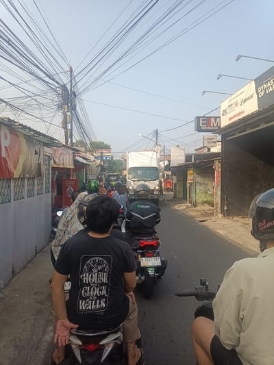 The image depicts a street scene in Depok, Indonesia, characterized by heavy traffic congestion. Multiple motorcycles are visible, with riders and passengers, some wearing helmets. A white truck is also part of the traffic flow. Along the sides of the street, there are various shops and businesses, some with signs and banners, including one for "EJM RETAIL PARTNER" and another advertising "LOST VAPE." Overhead, a dense network of electrical wires crisscrosses the sky, a common sight in urban Indonesian environments. The overall atmosphere suggests a typical busy day in a densely populated urban area of Depok.