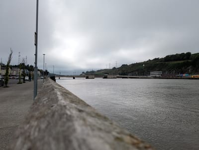 The image depicts a waterfront scene in Waterford, Ireland, under a cloudy, overcast sky, suggesting it is either early morning or late afternoon. The foreground is dominated by a weathered, wooden railing or wall, out of focus, creating a sense of depth. To the left, a paved walkway or promenade runs alongside the water, with several cars parked and some trees lining the edge. A lamppost stands prominently near the walkway.

Beyond the walkway, a calm body of water stretches out, reflecting the muted light of the sky. In the middle ground, a wide bridge spans the water, with vehicles visible crossing it. Further in the distance, a green, tree-covered hillside rises, dotted with some buildings and high-voltage electricity pylons. On the hillside, a small Irish flag is visible. The overall atmosphere is subdued and somewhat melancholic, fitting the cloudy weather. No specific activity or event is overtly taking place, giving the impression of a quiet, ordinary day. The scene captures a view of an urban waterfront integrated with the natural landscape.
