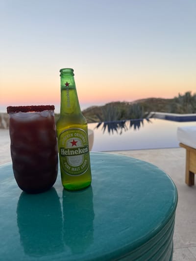 On a teal, circular glazed table, a frosted glass of a dark red beverage with a chili-salt rim and a condensation-covered green glass bottle of Heineken Original beer are positioned in the foreground. No individuals are visible. The background features an infinity pool reflecting the sky. Beyond the pool, arid hills with agave plants lead to the horizon where the ocean is visible under a sky transitioning from light blue to soft orange and pink, indicative of dusk. A portion of a modern outdoor lounge chair with a white cushion is partially visible on the far right. The scene depicts an outdoor patio or terrace, consistent with a resort environment in San José del Cabo, Mexico, overlooking the ocean and local desert landscape.