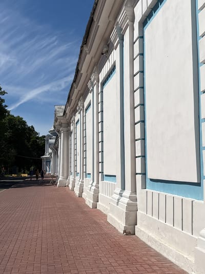 The image shows the facade of a white building with blue accents, featuring classical architectural elements like columns and ornate moldings. The building is situated alongside a red brick sidewalk. In the background, several people are walking on the sidewalk, and a line of trees borders a street. The sky above is blue with wispy white clouds. The architectural style and the distinctive blue trim are characteristic of the Narva Triumphal Arch complex in St. Petersburg, Russia.