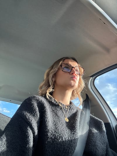A young adult female is depicted inside a vehicle, located in Chantemerle-les-Blés, France. She is seated and secured by a dark gray seatbelt extending diagonally across her chest. She wears a dark gray knit sweater and two layered gold chain necklaces, one featuring a circular medallion pendant. Her hair is blonde and styled in waves. She wears large, square, tortoiseshell-patterned eyeglasses that reflect ambient light. Her lips are pursed, and she directs her gaze upwards and to her right. Strong direct sunlight illuminates the right side of her face. The vehicle's interior features a light gray headliner. Through the windows, a bright blue sky with scattered white clouds is visible.