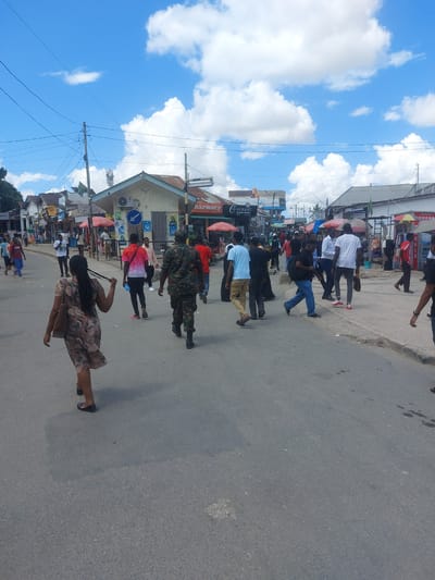 A medium-angle shot of a street scene in Dar es Salaam, Tanzania, shows a group of pedestrians walking along a paved road, presumably a busy commercial area with various shops and businesses lining the street. The sky is blue with scattered white clouds, suggesting a sunny day. Pedestrians walking in the same direction dominate the foreground. Shops and buildings can be seen in the background, and overhead electrical wires.
