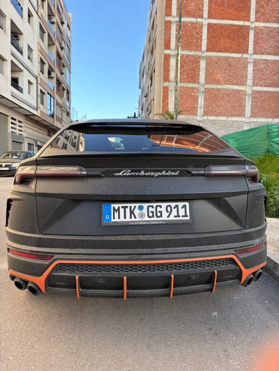 A matte black Lamborghini Urus, covered in a layer of dust, is parked facing away from the viewer on an asphalt surface in Meknes, Morocco. The vehicle displays a German license plate, "MTK GG 911", set in a frame that reads "AUTOPORT-L FRANKFURT.de". The rear bumper features orange accent lines and a diffuser with quad exhaust pipes. The "Lamborghini" emblem is visible on the tailgate.
To the immediate left of the vehicle, a dark sedan is parked. In the background, on the left, stands a multi-story residential building with light-colored facades, multiple balconies, and visible air conditioning units. On the right, another multi-story building is under construction, identifiable by its red brick walls and exposed concrete structural frame. Green construction netting is visible at the base of this building. The sky above is clear blue. No individuals or discernible actions are present in the scene.