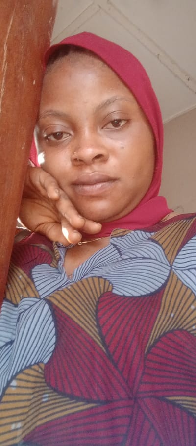 A female subject is depicted in a chest-up portrait within an indoor or semi-enclosed setting in the city of Adankolo, Nigeria. She possesses a medium brown complexion, dark eyes, and a neutral facial expression, looking directly at the viewer. A small metallic nose ring is visible on her left nostril. She is wearing a solid red headscarf that covers her hair and neck. Her attire includes a patterned garment featuring a V-neckline and a design composed of curved, striped fan-like shapes in red, gold, and light blue/grey, outlined with dark lines. A thin, gold-colored necklace with a small circular pendant is visible around her neck. Her left hand, with fingers slightly curled, rests against her chin and jawline. A vertical, light brown wooden structural element occupies the left side of the frame, partially obscuring her left arm and shoulder. The background in the upper right quadrant consists of a light-colored ceiling or wall with a discernible linear architectural detail. Illumination appears to be from a natural light source.