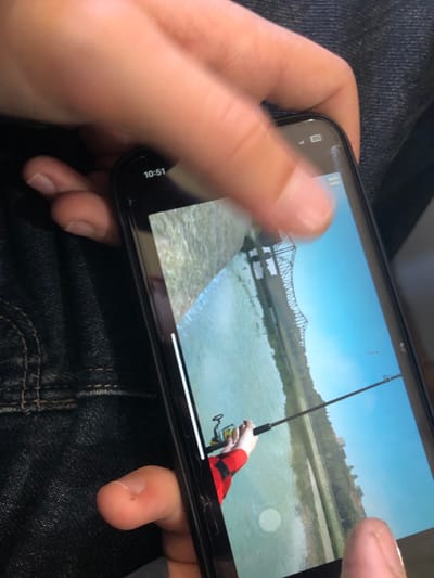 A close-up, high-angle photograph, captured in Ortabatumi, Georgia, depicts an individual's hands holding and interacting with a dark-colored smartphone against a blurred background of dark blue denim fabric. The smartphone's screen displays a landscape-oriented video, which features a person wearing a red and black checkered long-sleeved shirt, holding a fishing rod equipped with a yellow reel over a body of water. In the background of the video, a metal truss bridge spans the water, with green and tan land visible on both banks under a clear, bright blue sky. A fishing line extends from the rod into the water. The video playback interface includes a white scrubber bar along the left edge and a circular UI element in the lower right. The phone's status bar, partially visible at the top left, indicates the time "10:51" alongside cellular signal and Wi-Fi icons. The screen exhibits minor cracks or scratches, primarily near the top and right edges. The hands holding the device are of a light skin tone, with fingers positioned to operate the phone; one finger, likely a thumb, rests on the lower right portion of the screen, and another finger appears blurred above the top right, indicating motion or interaction. Fingernails are visible and appear unmanicured.