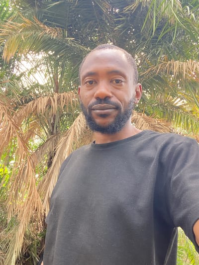 A man with dark skin and a full, dark beard is wearing a black t-shirt. He is standing under palm trees. The location is Sabon Tasha, Nigeria.