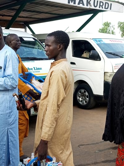 In Gombe, Nigeria, a group of men stand outdoors beneath a covered structure. In the foreground, a man wearing a tan, long-sleeved garment with intricate gold embroidery around the collar and cuffs holds a small, dark-colored spray bottle and a lighter-colored bottle in his right hand. He is also holding a blue plastic basket filled with various small items, including boxes and bottles, with his left hand. Beside him, to his left, stands another man in a pale blue, long-sleeved tunic. Further back, a third man in an orange garment is partially visible. Behind them, a white Toyota van with "TRP" and "TOYOTA" branding is parked. The van has tinted windows and a red stripe along its side. A sign with "MAKURI" is visible on the overhead structure. The ground appears to be paved.