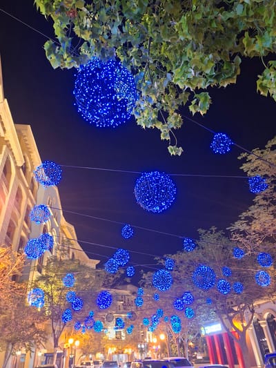 In Doha, Qatar, a street is adorned with numerous blue spherical lights hanging from wires strung between trees and buildings. The lights are dense clusters of small blue LEDs creating a festive, possibly holiday, atmosphere. The street is dark, indicating nighttime, and illuminated by the blue lights and some ambient streetlights and building lights. Trees with sparse foliage line the street, and building facades are visible in the background, some featuring arches and ornate architecture. The scene suggests a public space, possibly a commercial or residential area, decorated for an event.