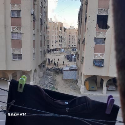 The image, taken from an elevated perspective, depicts a severely damaged urban residential area in Khan Yunis, Palestinian Territory. Multiple multi-story apartment buildings with light-colored facades and decorative window accents frame a central, unpaved area. Many windows on these buildings are broken, missing glass, or covered with makeshift materials; significant structural damage, including large open holes in upper floor walls, is evident on the rightmost building.
The ground level between the buildings is heavily covered with rubble, debris, and scattered refuse. Several makeshift tents or shelters constructed from tarpaulins are present in this central area. Approximately 10-15 individuals, consisting of adults and some children, are observed standing or walking in small groups amidst the debris.
In the immediate foreground, a dark garment hangs on a clothesline, secured by one green and one purple clothespin. Under the arched ground-floor supports of the building on the right, a vehicle is covered with a yellowish-green tarp, and a silver sedan is partially visible nearby. The sky above is partly cloudy.