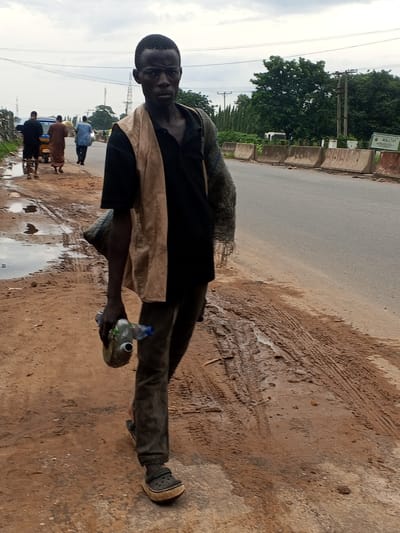 A man walks along the side of a road in Lokoja, Nigeria. He is carrying a grey bag on his left shoulder and a plastic bottle in his right hand. He is wearing a black shirt, a tan vest, dark pants, and black clogs. In the background, several other people walk on the road, and a yellow tricycle is visible. The road is paved, and the ground next to it is dirt and mud with tire tracks. Trees and utility poles are present in the distance. The sky is overcast.