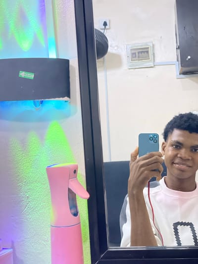 A young man is taking a selfie in a mirror. He is holding a blue iPhone. The man has short, dark hair and is wearing a white long-sleeved shirt with a black and white design. The background shows a room with an electrical panel, a fan, and a pink spray bottle. A strip light illuminates the area with green and blue light. The location appears to be a salon or barber shop. The city, Ado, Nigeria, is known due to the provided prompt.