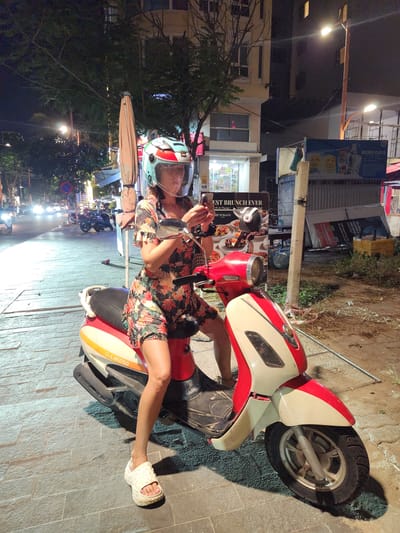 A person wearing a helmet and a floral print dress is sitting on a red and white scooter. They are holding a phone up to their ear, and their legs are crossed. The scooter has "BeLIKE50" written on its side. The scene appears to be a street at night, with buildings and trees visible in the background. There are streetlights and car headlights illuminating the area. Signs in Vietnamese are visible on buildings, one of which says "MENU CAFE". Another sign reads "EST BRUNCH EVER BRUNCH&CO.". The ground is paved with stone tiles. The overall mood is casual, possibly a break during a nighttime outing.