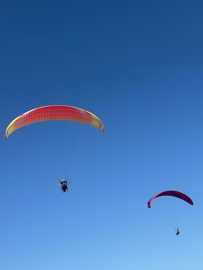 The image depicts two paragliders against a clear, vibrant blue sky. The paraglider on the left is larger and more prominent, featuring a red and yellow wing. Two individuals are visible under this wing, appearing to be a tandem paragliding setup. The second paraglider, smaller and further to the right, has a pink and black wing and also carries a single individual. The scene is set in Kuta Selatan, Indonesia, an area known for its suitability for paragliding due to its coastal geography and favorable wind conditions. The image captures a moment of recreational activity and airborne freedom in a tropical environment.