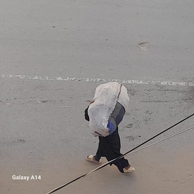 An overhead, medium shot depicts a person walking on a concrete surface, likely a street. The person is carrying a large, white sack on their back. They are wearing dark clothing and light-colored sandals. Power lines are visible in the upper right corner of the frame. A worn white line is present on the concrete to the left of the person. "Galaxy A14" is printed in the lower right corner. The location is presumed to be the city of Khan Yunis, Palestinian Territory, based on the prompt.
