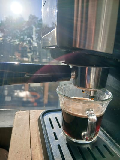 A close-up image captures a coffee machine in the midst of brewing, with a glass mug partially filled with dark coffee. The clear glass mug, featuring a visible handle on its right side, rests on a metallic drip tray with slotted perforations. A thin layer of light-brown crema floats atop the coffee in the mug.

From the left, the prominent brew head of the coffee machine is visible, a cylindrical component made of shiny silver and dark grey metal. Light glares off its polished surface. Extending downwards from the brew head is a dark, elongated handle, likely part of a portafilter, which also catches reflections.

The coffee machine is situated on a light-colored wooden surface, visible towards the bottom and right edges of the frame. The background, significantly out of focus, shows bright sunlight and reflections of outdoor scenery, including what appears to be trees and vague structural forms, indicating a window or an outdoor setting. Intense sun glare and lens flare are noticeable, especially on the metallic parts of the machine and in the bright background, signifying a sunny daytime. No discernible text is visible within the frame. The scene is set in San Fernando, Argentina.