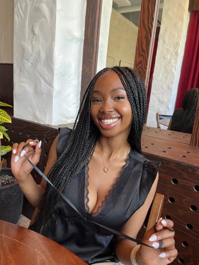 A young Black woman with braided hair, wearing a black satin top with lace trim and a gold necklace, smiles at the camera. She is holding a thin black ribbon in her hands, which are adorned with white nail polish. The background appears to be an indoor setting, possibly a restaurant or cafe, with textured white walls and wooden accents. A mirror reflects another person, also with braided hair, seated at a table. The scene is well-lit, with natural light contributing to the overall brightness. The woman is seated at a wooden table, and to her left, a potted plant is visible. The location is not Kazan, Russia.