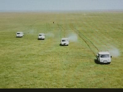 Four white recreational vehicles are driving across a grassy plain under a bright, clear sky. The vehicles are spaced out, with the closest one to the viewer on the right side of the image. Each vehicle appears to be kicking up a small amount of dust or mist from the ground as it moves. In the distance, a single dark figure, possibly a person, stands on the plain, a tiny speck against the vast green landscape. The scene suggests a journey or exploration in an open, undeveloped area. The time of day appears to be daytime, given the bright sky and shadows cast by the vehicles. The location is implied to be Shymkent, Kazakhstan, which is known for its vast plains and open spaces. There is no visible text in the image.