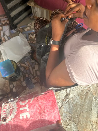 A person's hands are shown in the foreground, sewing a piece of dark fabric with white thread, using a needle and thimble. The person is wearing a light pink t-shirt and a watch on their left wrist. In the background, a patterned fabric and a pair of blue-handled scissors are visible. To the left, a white plastic bag and a blue plastic container are on the ground, which is tiled with colorful mosaic patterns. The scene appears to be outdoors, possibly in a courtyard or on a patio, with sunlight casting shadows. The style of the tiles and the general ambiance suggest a location in Badagry, Nigeria.