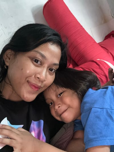 A selfie captures a woman and a young child lying down. The woman, with dark hair and tan skin, smiles directly at the camera. The child, slightly above and behind her, has dark hair and a playful expression. The child wears a blue shirt. A red pillow rests behind them. The woman wears a black top. The image is taken at a close angle, creating a personal and intimate feel. The floor is visible in the background.