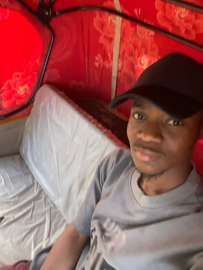 A single Black male is seated within the passenger compartment of a motorized tricycle (keke napep) in Minna, Nigeria. The individual wears a black baseball cap and a grey short-sleeved shirt, looking directly towards the viewer. The vehicle's interior is lined with red fabric featuring a lighter floral pattern. A white, plastic-covered seat is positioned to the left. Black metal frame elements are visible against the red interior.