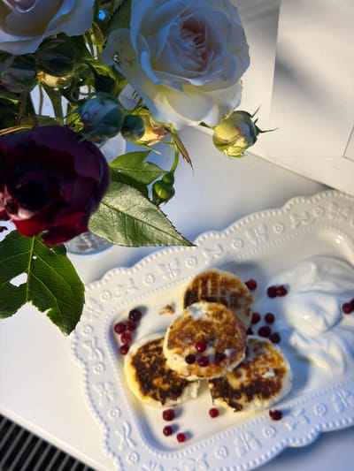 The image shows a still life arrangement. In the foreground, there is a bouquet of roses, including white and dark red blooms. The roses are in a vase (partially visible). Adjacent to the flowers is a white, decorative plate containing several small, round pancakes or fritters topped with red berries. The plate also has a molded design of cherubs. Scattered around the pancakes are more of the same red berries. The scene is set on a white surface, possibly a table, with a blurred striped pattern visible near the bottom right corner of the image. A white box or frame is present in the upper left corner. It's difficult to pinpoint the precise location in Rossiyskiy, Russia, without further context or landmarks in the image.