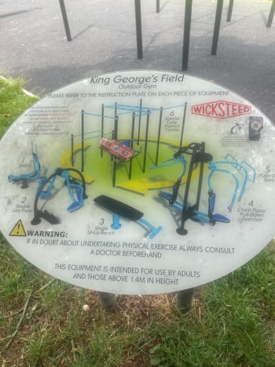 This image shows an outdoor gym located in King George's Field, Chessington, KT9 1TR, London, United Kingdom. The gym equipment is installed on a circular instruction plate made of a clear, durable material, possibly Perspex, which is placed on a circular concrete base. The plate displays information about the equipment, including numbers, exercise names, and safety warnings.
The equipment visible includes a "Skier," a "Double Leg Press," a "Single Sit-Up Bench," a "Chest Press/Pull-Down Exerciser," and "Push-Up Bars." The equipment is predominantly blue and black, with some yellow accents visible on the instruction plate itself. There is also a "Special Calisthenics Centre" indicated on the plate, though the specific equipment for this is not clearly discernible.
The instruction plate features a prominent "WICKSTEED" logo, with "SINCE 1918" visible. It also includes a QR code for users to scan for exercise videos and contact information for further information, fault reporting, and emergencies. A significant warning states, "IF IN DOUBT ABOUT UNDERTAKING PHYSICAL EXERCISE ALWAYS CONSULT A DOCTOR BEFOREHAND" and "THIS EQUIPMENT IS INTENDED FOR USE BY ADULTS AND THOSE ABOVE 1.4M IN HEIGHT."
The scene is set outdoors, with the instruction plate and gym equipment surrounded by green grass and some scattered twigs and branches, suggesting a park or recreational area. The lighting suggests it is daytime. No people are visible in the image, and the overall impression is of a well-maintained public facility for outdoor exercise.