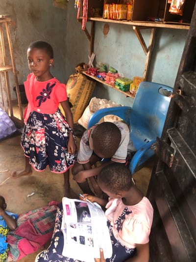 The scene depicts an interior space in Ukwu-Abwa, Nigeria, functioning as a small, rudimentary shop or dwelling. Four children are present. A young girl, barefoot and wearing a coral top with a floral skirt, stands centrally, facing slightly towards the viewer. To her right, two children are seated on the concrete floor; one, in a grey and white striped shirt, leans forward, while a girl in a light pink top and floral skirt holds an open textbook containing images and text, actively writing with a pen. A fourth child is partially visible lying on a plaid blanket in the lower-left corner. The light blue walls are unpainted and show wear. Rustic wooden shelves, supported by planks, are mounted on the back wall, displaying various packaged goods such as sweets, snacks, and sachets. Large woven sacks are stacked on the floor beneath the shelves. An overturned blue plastic chair is visible on the right, near a dark, possibly metal, frame or door structure. A small, pale lizard is on the floor near the feet of the standing girl.
