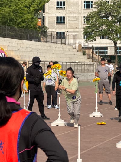 A group of people, dressed in casual clothing and athletic wear, are positioned along a lane on a track and field in Zhongtun, China. They are holding sunflowers. The track surface appears to be a brown or reddish-brown color, and it is marked with white lines. White lane dividers are positioned along the track. In the background, there is a multi-story building with rectangular windows. Trees are visible on either side of the building. A concrete structure, possibly bleachers, runs along the side of the track near the building. The weather seems sunny.