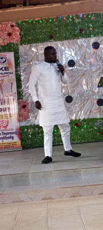 The image displays an indoor scene within a building in Akwanga, Nigeria, showing a male individual on a raised platform. The man, an adult with dark skin and short dark hair, is positioned slightly right of the frame's center. He wears a white long-sleeved tunic, white trousers, and black shoes. A gold-colored ring is visible on his right index finger. He holds a blue and black microphone to his mouth with his right hand, his left foot slightly raised, suggesting movement.
The background consists of layered decorations. The uppermost section features a horizontal panel of artificial green foliage, interspersed with small red, green, and blue LED lights. Below this, a silver-colored, crinkled, reflective material forms a backdrop, on which several circular black objects with horizontal stripes are affixed. Clusters of artificial pink and red flowers are placed on the green foliage panel and the silver backdrop.
To the left, a tall, vertical banner is visible, displaying a "BAPTIST CONVENTION" logo, "KE CHURCH (LIGHT)", an illustration of a white armchair, and the text "UNLIMITED FAVOUR 2023". Further text includes "Za Sami Tagomashi" and a Bible verse reference "Prov 3:1 NLT". The man stands on a light-colored tiled platform, which transitions via a step to a lower, similarly tiled floor in the foreground. The top edge of the image reveals a brown structural element, possibly a ceiling beam.