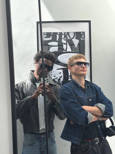 The image shows two young men posing for a mirror selfie in what appears to be an indoor setting. The man on the left, with dark, curly hair and sunglasses, is wearing a black leather jacket over a white t-shirt and blue jeans. He is holding a smartphone and taking the picture. The man on the right, with blonde, curly hair and sunglasses, is wearing a denim jacket over a black t-shirt and dark jeans. He has his arms crossed. In the background, a framed black and white artwork is visible on a white wall. The reflection in the mirror suggests a minimalist aesthetic. There are no visible clues to suggest the location is in London, United Kingdom.