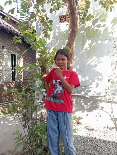 A young Indonesian girl, likely of elementary school age, stands outdoors in what appears to be a residential area of Baron, Indonesia. She is wearing a red t-shirt with a graphic design and light blue denim jeans. Her hair is dark and pulled back, and she has a light complexion. She is positioned near a tree with a rough, brown trunk and is leaning slightly to her right. Her left arm is bent, with her hand near her chin, and her right hand is raised, with her thumb and index finger extended.
Behind the girl is a wall, partially obscured by foliage and shadows from a nearby tree. The wall has a rough, textured surface, possibly plaster or stucco, and is a pale, off-white color. A decorative, circular vent is visible on the wall. To the girl's left, a section of a brick building is visible, with a wooden window frame and shutters. The building has a tiled roof. The ground in front of the girl is covered with small, gray gravel and some scattered red bricks. In the background, further foliage and what might be a fence or railing are visible. The overall impression is a sunny, outdoor scene in a rural or suburban Indonesian setting.