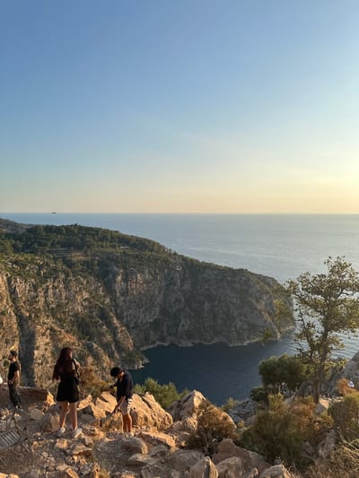 In Fethiye, Turkey, three individuals are seen on a rocky cliff overlooking a vast expanse of the sea. The scene is bathed in the warm, golden light of late afternoon or early evening, suggesting a sunset or sunrise. The sky transitions from a pale blue near the horizon to a hazy yellow, indicating either the setting sun or atmospheric haze.
The foreground is dominated by the rugged texture of the rocky cliff, with sparse vegetation clinging to its surface. The sea stretches out to the horizon, appearing calm and a deep blue. In the distance, a narrow, dark inlet or canyon carved into the coastline is visible, adding depth and drama to the landscape. The greenery on the slopes of the cliff and the distant land suggests a Mediterranean or coastal environment.
Two of the individuals are positioned near the edge of the cliff, appearing to be looking out at the view. One person, wearing a dark t-shirt and light shorts, is bending over, perhaps taking a photograph or examining something on the ground. Another individual, wearing a black dress, stands closer to the cliff edge, also facing the sea. A third person, wearing a black t-shirt and dark pants, is seated or reclining further down the slope. The overall atmosphere appears relaxed and contemplative, with the individuals seemingly enjoying the scenic vista. No text is visible in the image.