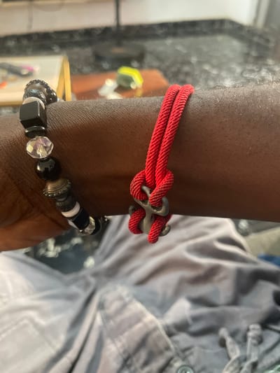The image shows a close-up of a person's arm wearing two bracelets. One bracelet is made of black and white beads, with a large faceted crystal bead and a square black bead. The other bracelet is a simple red cord with a metallic anchor charm tied into the knot. The arm is dark-skinned, and the background is blurred, showing hints of furniture and a textured surface that could be a desk or table. The person appears to be wearing a gray, possibly wrinkled, fabric garment. There are no discernible city-specific features or indications of Abuja, Nigeria within the image itself.