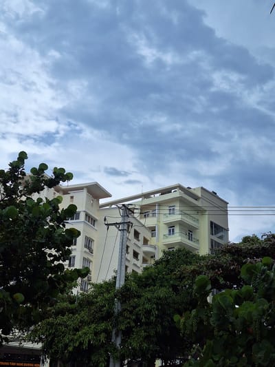 The image shows a multi-story, light yellow building with balconies and large windows, partially obscured by lush green trees and foliage in the foreground. A utility pole with electrical wires is visible to the right of the building. The sky above is overcast with gray clouds. A sign is partially visible at the bottom left of the image, appearing to be in Vietnamese. The building's architectural style and the presence of tropical vegetation suggest a location in a coastal city. This scene is consistent with the environment found in Nha Trang, Vietnam, a well-known beach resort city.