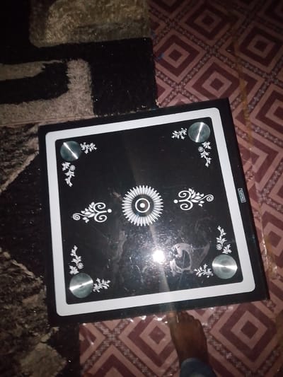 A square, black carrom board with white decorative edges sits on a patterned maroon and white floor. Four silver circular pieces are embedded at the corners of the board, and a central circular design with radiating lines is present in the middle of the board. A person's foot is visible at the bottom right of the image, partially on the board. The background consists of a dark, textured rug and the patterned floor. The scene is likely indoors in a residential setting in Jauro Yinu, Nigeria.