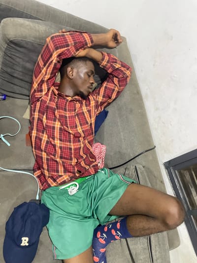 An adult male is asleep on a gray sofa in an indoor setting, possibly an apartment or living room. He is wearing a red and orange plaid long-sleeved shirt, green shorts with white drawstrings, and blue socks with a cherry pattern. His arms are crossed above his head, and he appears to be sweating. To his left, on the sofa, is a dark blue baseball cap with a white "R" embroidered on it. Several electrical cords are visible on the sofa, including a light blue one with a white connector and a black cord. A small plastic bottle and a red and white patterned cloth are also on the sofa. The background includes a white wall and a dark-framed window or door. The scene is not specifically indicative of Abuja, Nigeria, but could be a domestic setting within the city.