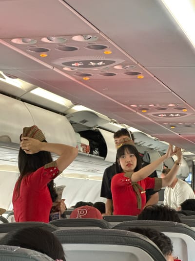 In an airplane cabin, a flight attendant in a red uniform with a checkered collar and buttons on the sleeves is the primary subject. She is gesturing with both hands, her arms raised, and her face is turned towards the camera with a slight smile. Her dark hair frames her face. She is wearing a checkered headband.
Behind her, other passengers are visible. One man, wearing a white t-shirt, is seen in the background. Another passenger in the foreground, also in a red uniform similar to the flight attendant's, is visible from the waist down, holding what appears to be a brochure or pamphlet in their hand. Other passengers are seated in the rows of airplane seats, some with dark hair. One passenger in the foreground is wearing a red baseball cap with a white logo.
The scene is set within an airplane, indicated by the rows of seats, overhead compartments with air vents and lights, and the general interior of the cabin. The overhead lights are on, suggesting it is likely daytime or that the cabin is artificially lit. The image has a slightly blurred quality, and the perspective is from a low angle, looking up towards the flight attendant. The overall atmosphere seems casual and potentially posed, given the flight attendant's direct gaze and gesture towards the camera. There is no visible weather information. The location cue is Kuta, Indonesia.