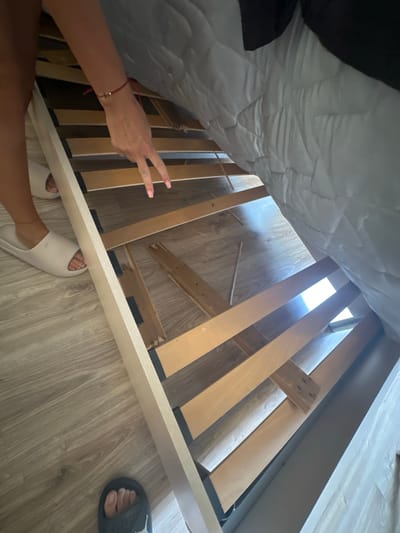 The image shows a damaged wooden bed frame with a white mattress on top. A person's legs and feet are visible, one wearing a white slide sandal and the other a black slide sandal. The person is reaching under the bed frame with their hand, which is adorned with a red string bracelet with a gold cross charm. The wooden slats of the bed frame are broken in several places. The floor appears to be light-colored laminate wood flooring. The scene is indoors, under a bed. No specific location in Samokov, Bulgaria can be determined from the image.