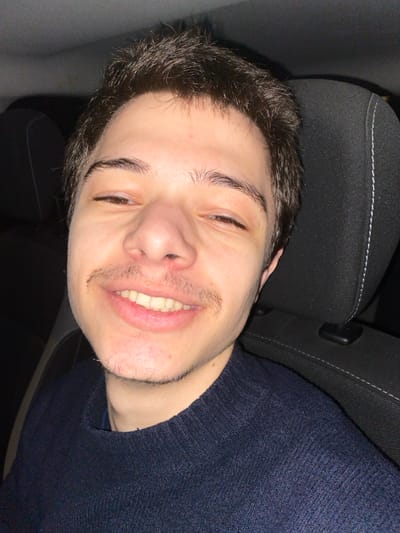 A young man with short dark hair and light skin smiles slightly, his eyes squinting. He has a mustache and a hint of a beard. He's wearing a dark blue sweater, seemingly in a car. The headrest behind him is black fabric with a white stitched seam. The image is rotated, making the headrest appear on the left side. The lighting is dim, creating shadows on the face.