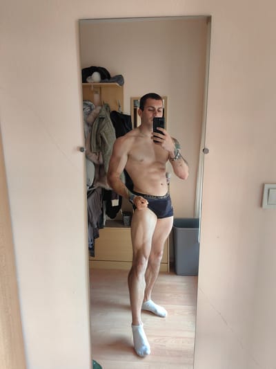 A man is taking a mirror selfie in what appears to be a bedroom or dressing area. He is shirtless and wearing black underwear and white socks. He is standing with his left leg slightly forward, and he is flexing his right arm. In the reflection, a closet with clothes is visible, as well as a trash can and part of a wall. The floor is made of light-colored wood. The overall lighting suggests it is daytime. There are no indications of the city of Varna, Bulgaria in the image.