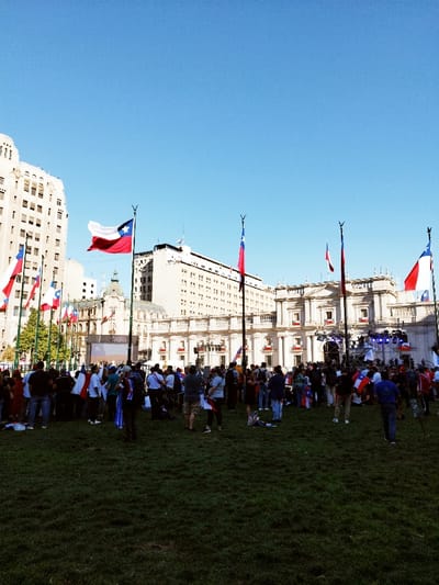 The image captures an expansive outdoor gathering on a manicured grassy lawn within Plaza de la Ciudadanía, situated in front of La Moneda Palace in Santiago, Chile. The scene unfolds under a clear, bright blue sky during daytime. La Moneda Palace, a prominent neoclassical structure with multiple columns and balconies, dominates the background, adorned with numerous Chilean flags draped from its windows and balconies. A large stage equipped with lighting fixtures is erected directly in front of the palace's central entrance. To the left, a tall, light-colored multi-story urban building with numerous windows is visible. Several tall flagpoles are dispersed across the square, each displaying a large Chilean flag billowing in the wind. Hundreds of individuals, diverse in age and attire, populate the grassy area, standing, observing, and moving. Many attendees are holding small Chilean flags or wearing patriotic clothing in the national colors. A large outdoor screen, positioned to the left of the main stage, displays imagery consistent with the Chilean flag. The assembly appears to be a public event or celebration.