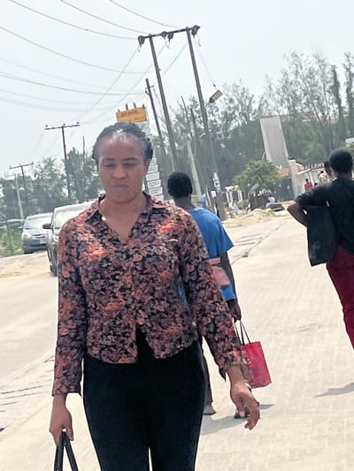 The scene depicts a daytime urban street in Lekki, Nigeria. In the foreground, an adult female with dark, braided hair walks towards the viewer's left, looking slightly downward. She wears a long-sleeved collared shirt with a black base and orange/beige floral patterns, paired with black trousers. Her left hand holds a dark object, possibly a bag.

In the mid-ground, behind the primary subject, an adult male walks away from the viewer, attired in a blue t-shirt and light-colored trousers, carrying a red shopping bag in his right hand. Further to the right, another adult individual is partially visible, wearing a dark top and red pants, with their left hand positioned on their hip. A light-colored sedan is visible on the street to the left.

The environment features a light-colored paved road surface. Multiple wooden utility poles supporting numerous overhead power and communication lines dominate the mid-background. A prominent yellow sign with red "DHL" lettering is affixed to one of these utility poles, indicating proximity to a DHL service point. Another sign, partially visible as "GALLERY," is located further down the street to the right. Trees and vegetation line the background, and the sky is bright and overcast. The overall setting is a commercial or mixed-use street within Lekki, Nigeria.