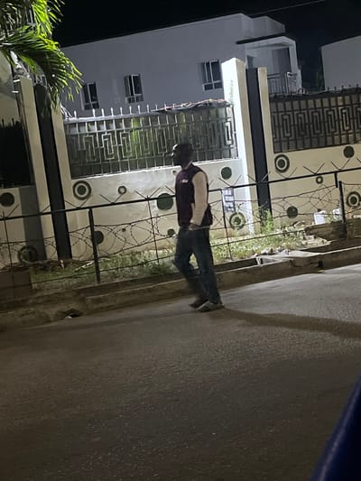 The scene depicts a street in the city of Lagos, Nigeria, at night. A lone male individual is walking from right to left across the asphalt road, positioned roughly in the center of the frame. He wears a dark-colored cap, a maroon and light-colored letterman-style jacket, and dark blue jeans. His gaze appears directed forward.

Behind the individual, a residential property is visible. It features a light-colored, possibly white or off-white, two-story building with multiple dark-framed windows. The property is secured by a perimeter fence system: an upper dark metal gate with intricate geometric patterns, mounted on a low wall. Below this, a dark metal fence spans the foreground, topped with multiple strands of coiled barbed wire. The wall supporting the main gate is light-colored and decorated with evenly spaced dark circular patterns. A strip of unkempt green grass and sparse vegetation separates the fence from the concrete curb and the street. To the far left, a portion of a palm tree with bright green fronds is visible, illuminated by an unseen light source. The street surface is dark, indicating a nighttime setting with artificial lighting casting a shadow of the walking man to his left. A small drain opening is visible in the curb line.