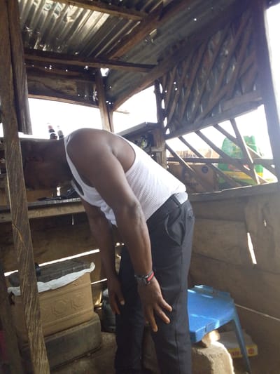 The image depicts an individual in a lean-to structure in Lokoja, Nigeria. The person, clad in a white tank top and dark trousers, is bending over. The structure appears to be constructed from wood and corrugated metal. A blue plastic stool and a stack of containers are visible within the space. Light streams through openings in the walls and roof. There are items such as bottles and boxes visible through the structure's openings, situated on shelves.
