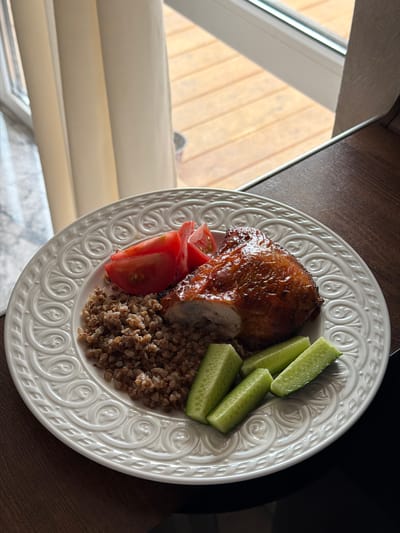 The photograph depicts a white plate containing a meal consisting of a piece of cooked chicken with a slice removed, buckwheat, sliced tomatoes, and cucumber sticks. The plate rests on a dark wooden surface near a window. The background visible through the window shows a wooden structure or fence. The image appears to have been taken indoors, likely in a dining or kitchen area. The plate itself is decorated with a pattern of repeating scroll designs. The location may be in Rossiyskiy, Russia, but there is no contextual evidence to confirm the city.