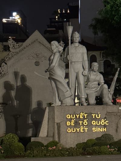 This is a nighttime photograph taken in Hanoi, Vietnam, showcasing a monument with several statues of people. The monument features a stone inscription with golden lettering. The statues depict individuals in what appears to be a historical or patriotic scene, possibly related to defense or struggle, given the presence of what looks like a rifle. One statue of a man holds a long, pole-like object, while another man behind him appears to be aiming a rifle. A female figure is positioned lower down, seemingly in a protective or supportive stance.
The setting is dimly lit, suggesting it is nighttime. The monument is situated outdoors, with greenery and bushes visible to the right. Shadows cast by the statues and possibly other elements are prominent on a textured, possibly carved, wall behind them. The overall mood is solemn and commemorative.
The visible text on the stone monument is in Vietnamese: "QUYẾT TỨ QUỐC ĐỂ TÔ QUỐC QUYẾT SINH". This translates to "Determination for the Nation, to sacrifice for the Nation." The golden lettering is clear and stands out against the stone. In the background, faint lights suggest the presence of buildings or streetlights, indicating an urban environment.