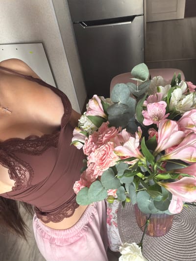 In Shushary, Russia, a person wearing a brown lace-trimmed camisole and pink pajama bottoms is positioned near a bouquet of pink and white flowers. The scene appears to be indoors, possibly a home, with a refrigerator visible in the background. The camera is angled downwards, focusing on the upper body of the individual and the floral arrangement.