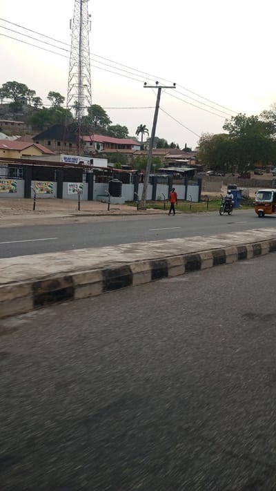 This image captures a street scene in Lokoja, Nigeria, likely in the late afternoon or early evening under a bright, slightly overcast sky. The foreground shows a multi-lane asphalt road with a concrete median featuring black and white striped curbs.
On the right side of the road, a person wearing an orange shirt walks along the shoulder. Further down the road, a motorcyclist in a white helmet is visible, and a yellow and blue tricycle (known as Keke Napep in Nigeria) is also present.
Beyond the road and median, there's a dark fence with multiple advertising boards. Text visible on these boards includes "DREY'S" (repeated) and "DAYLIGHT PERFECT LOGICS INVESTMENT LTD". Behind the fence, several buildings are visible, including what appears to be a commercial or residential structure with a black water tank placed on a stand. White-and-black striped street light poles are also spaced along the roadside.
In the mid-ground, a tall communication tower stands prominently, with power lines running across the frame, supported by a wooden utility pole. The background features rolling hills covered in trees and scattered buildings, some with red roofs, adding to the urban-rural blend of the landscape. A distinct palm tree is also visible against the sky. The overall impression is that of a typical street environment in a Nigerian town.