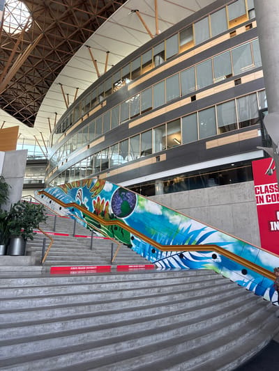 The image shows an interior view of a modern building with a curved facade and a large wooden, geometrically complex ceiling structure. In the foreground, a wide, curved concrete staircase with red risers bearing the text "SIMON FRASER UNIVERSITY" ascends. The handrail of the staircase is adorned with a vibrant mural depicting a stylized planet Earth with hands reaching out, clouds, tropical leaves, and possibly a ship, painted in blues, greens, and yellows. The building's interior features a grey concrete pillar on the right and a large red banner on the far right with white text, partially obscured, reading "CLASS IN CON..." and "IN". To the left of the stairs, a large metallic planter holds green foliage. The overall environment suggests an institutional or public space, likely a university campus. Based on the text on the risers, the location is Simon Fraser University. The specific campus of Simon Fraser University in Surrey, Canada is the Surrey campus.